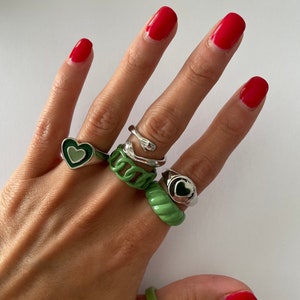 May include: A hand wearing five rings. The rings are silver and green. Two of the rings are heart-shaped. One ring is a snake design. Two rings are green and have a ridged design.