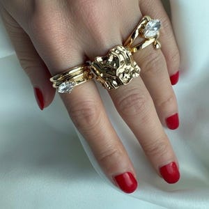 Può includere: Primo piano di una mano ornata da diversi anelli d'oro. Uno presenta un design strutturato e organico, mentre altri hanno pietre chiare a forma di goccia. Le unghie sono dipinte di un rosso vibrante, creando un contrasto con l'oro.