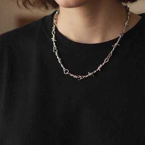 Könnte beinhalten: Silberfarbene Halskette mit einem Stacheldraht-Design. Die Halskette besteht aus Metall und weist eine Reihe kleiner, spitzer Widerhaken entlang der Kette auf. Die Halskette wird um den Hals getragen und liegt auf einem schwarzen Oberteil.