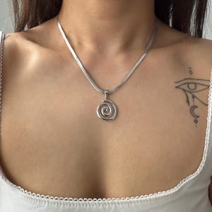 Peut inclure: Un collier argenté avec une chaîne plate en chevrons et un pendentif en spirale. Le pendentif présente un motif ajouré. Le collier est porté avec un haut blanc.