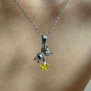 May include: Silver-toned necklace featuring a small bear pendant holding a yellow star. The bear pendant has a detailed design, and the star has a textured appearance. The necklace is worn against a neutral background.
