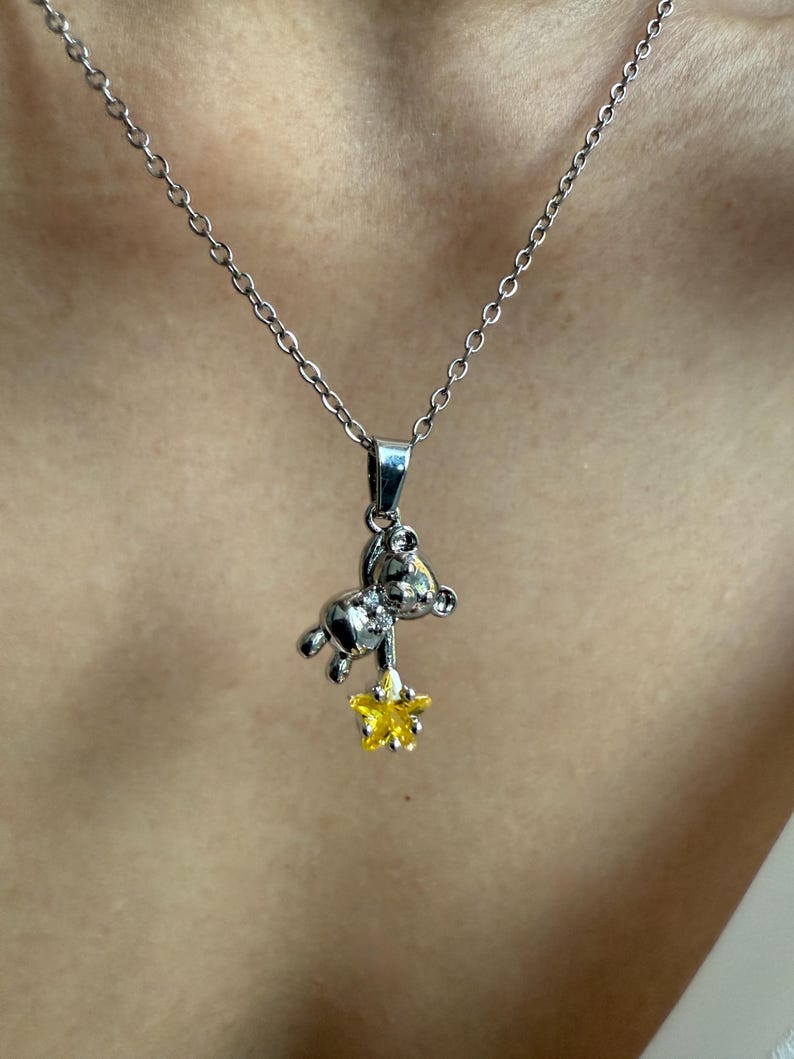 May include: Silver-toned necklace featuring a small bear pendant holding a yellow star. The bear pendant has a detailed design, and the star has a textured appearance. The necklace is worn against a neutral background.