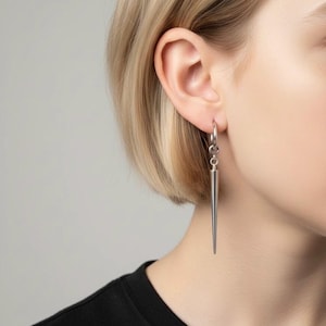 Könnte beinhalten: Ein silberner Ohrring mit einem langen, spitz zulaufenden, konischen Design. Der Ohrring hängt an einem kleinen Reifen. Die Person, die ihn trägt, hat kurze blonde Haare und ein schwarzes Oberteil. Der Ohrring ist etwa 5 cm lang.