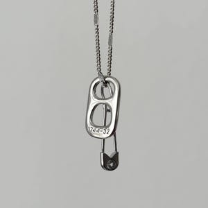 Puede incluir: Un collar plateado con un colgante de anilla de lata y un imperdible. La anilla tiene grabado el número "1744-32". El collar cuelga de una delicada cadena sobre un fondo neutro.