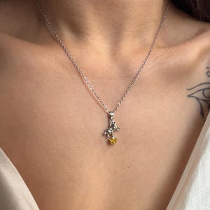 May include: A silver necklace with a small pendant featuring a figure riding a horse and a yellow gemstone star. The necklace is worn against a person's skin and a white top.
