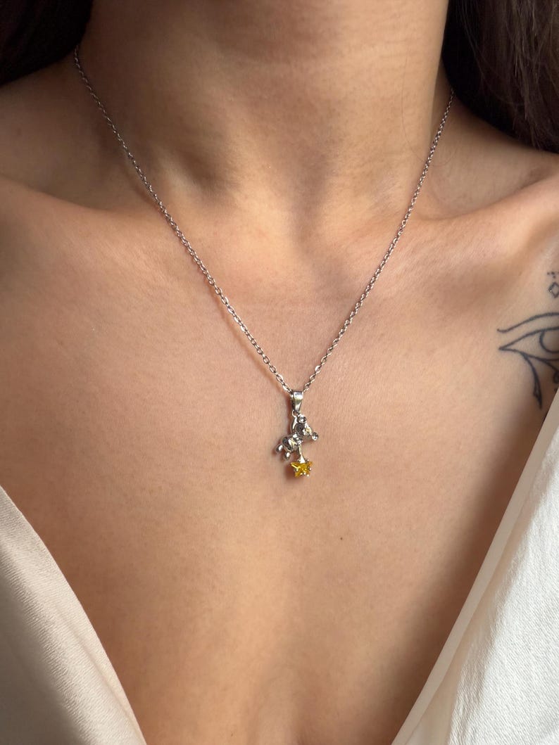 May include: A silver necklace with a small pendant featuring a figure riding a horse and a yellow gemstone star. The necklace is worn against a person's skin and a white top.