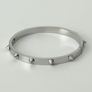 Pode incluir: Uma pulseira de bracelete articulada prateada com múltiplas pontas cônicas espaçadas uniformemente no exterior. A pulseira é feita de um material metálico brilhante e é exibida em uma superfície branca. É uma joia.