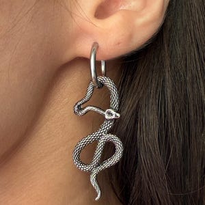 May include: A silver-toned hoop earring featuring a dangling, textured snake charm. The snake has a detailed scale pattern across its body and a small, clear stone set into its head, hanging vertically from a smooth, circular metal hoop.