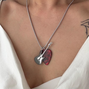 Puede incluir: Collar plateado con un colgante que presenta una guitarra en miniatura y una púa. La guitarra es gris y blanca, y la púa es roja con un patrón oscuro. El collar se muestra sobre una tela blanca.