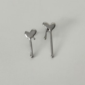 May include: A pair of silver-toned metal earrings featuring a heart shape with a liquid-like dripping effect. Each earring has a smooth, reflective surface with a single elongated drop extending downward from the base of the heart design.