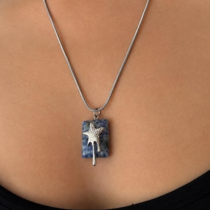 May include: A silver-toned snake chain necklace featuring a rectangular pendant made of mottled blue and grey stone. Attached to the front of the stone is a silver-toned metal charm shaped like a star with a dripping liquid effect.