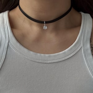 Könnte beinhalten: Ein schwarzes Choker-Halsband mit einem kleinen, runden, klaren Edelsteinanhänger. Die Halskette besteht aus einem dünnen, dunklen Material und liegt eng am Hals an. Der Anhänger hängt an einer silberfarbenen Öse.