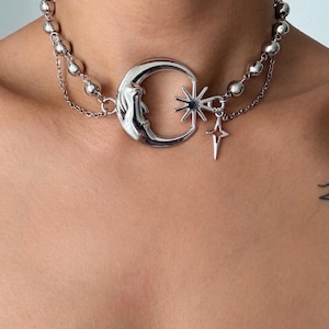May include: A silver-toned choker necklace featuring a row of polished spherical beads and a delicate chain. The centrepiece is a crescent moon charm with a face, accompanied by a multi-pointed star charm and a smaller four-pointed star pendant.