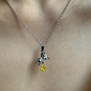 May include: Silver-toned necklace featuring a small bear pendant holding a yellow star. The bear pendant has a detailed design, and the star has a textured appearance. The necklace is displayed against a skin-toned background.