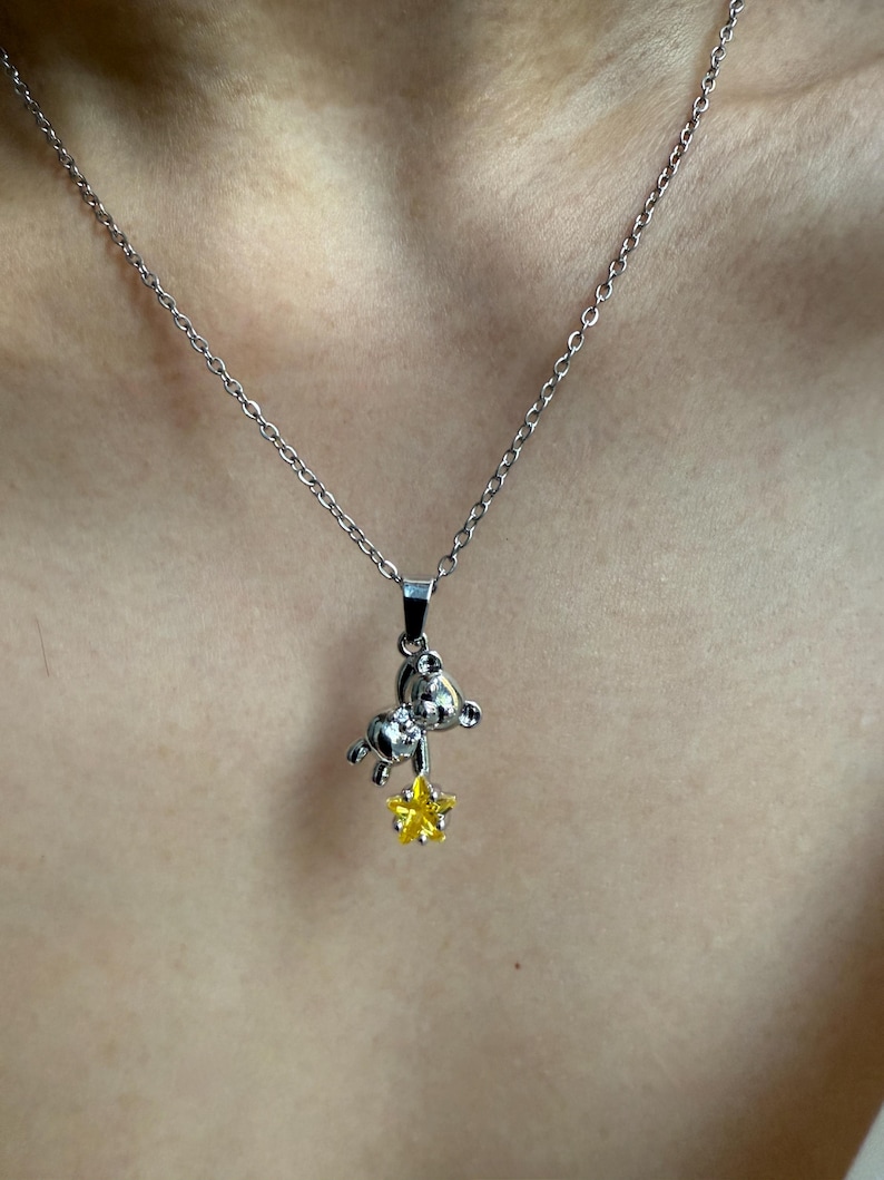 May include: Silver-toned necklace featuring a small bear pendant holding a yellow star. The bear pendant has a detailed design, and the star has a textured appearance. The necklace is displayed against a skin-toned background.