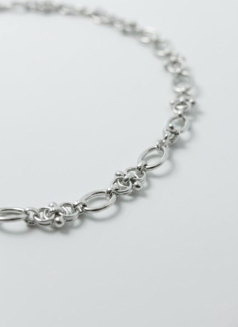 Macro photography of a silver-tone gothic necklace focusing on the unique "pierced" link structure and the polished, tarnish-resistant surface of the steel.