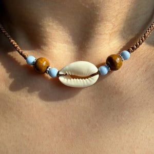 Peut inclure: Un collier fait à la main avec un pendentif coquillage crème, des perles en bois et des perles bleu clair. Le collier est monté sur un cordon marron. Un accessoire inspiré de la plage.