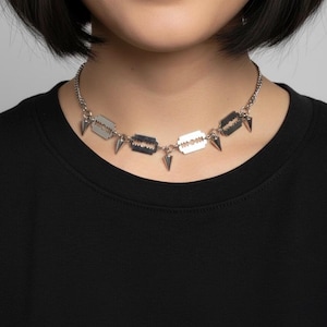 Könnte beinhalten: Silberfarbene Halskette mit rasiermesser- und spitzförmigen Anhängern. Die Halskette wird über einem schwarzen Oberteil getragen, was einen auffälligen Kontrast erzeugt. Das kantige Design ist ein auffälliges Statement-Piece.