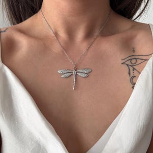 Könnte beinhalten: Eine silberne Libellen-Anhänger-Halskette. Der Anhänger hat detaillierte Flügel mit kleinen, funkelnden Akzenten. Die Libelle hängt an einer zarten Silberkette und schafft ein elegantes, von der Natur inspiriertes Accessoire.