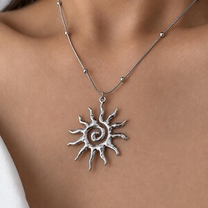 Puede incluir: Collar plateado con colgante de sol. El colgante tiene un diseño de sol texturizado y estilizado con un centro en espiral y rayos radiantes. La cadena incluye pequeñas cuentas redondas. Una joya.
