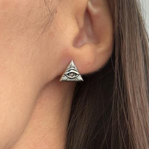 Op de afbeelding: Een zilverkleurige oorsteker in de vorm van een driehoek, voorzien van een gegraveerd oog in het midden met stralende lijnen. Het ontwerp bevat een klein, donker cirkelvormig accent in de pupil op een getextureerd metalen oppervlak.