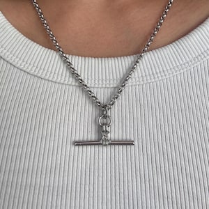 May include: A silver-toned necklace featuring a rolo chain and a toggle clasp pendant. The pendant consists of a horizontal bar attached to a circular ring, creating a clean, metallic design that rests against a white ribbed fabric background.