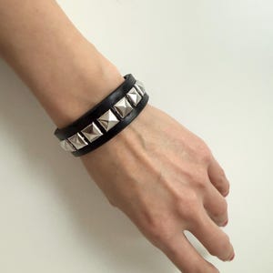 May include: Black leather bracelet with silver pyramid studs. The bracelet is a wide cuff style, with a row of square studs. The bracelet is worn on a wrist.