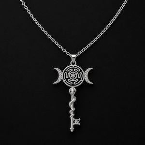 Op de afbeelding: Een zilverkleurige ketting met een hanger in de vorm van een sleutel. De kop van de sleutel heeft een cirkelvormig labyrintontwerp, geflankeerd door twee maansikkels bezet met kleine heldere steentjes. Een slang kronkelt om de schacht.