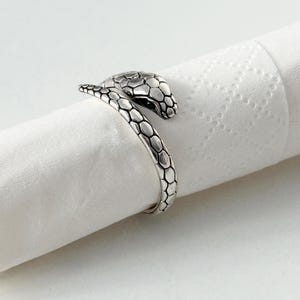 Peut inclure: Une bague en métal argenté en forme de serpent enroulé. La bague présente un motif d'écailles texturé et détaillé sur toute sa surface, avec la tête du serpent reposant légèrement au-dessus de la queue, créant un design de bande ouverte.