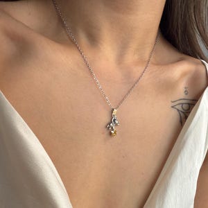 May include: A silver necklace with a small pendant featuring a detailed design with gold and yellow accents. The necklace is worn on a person's chest, against a white top.