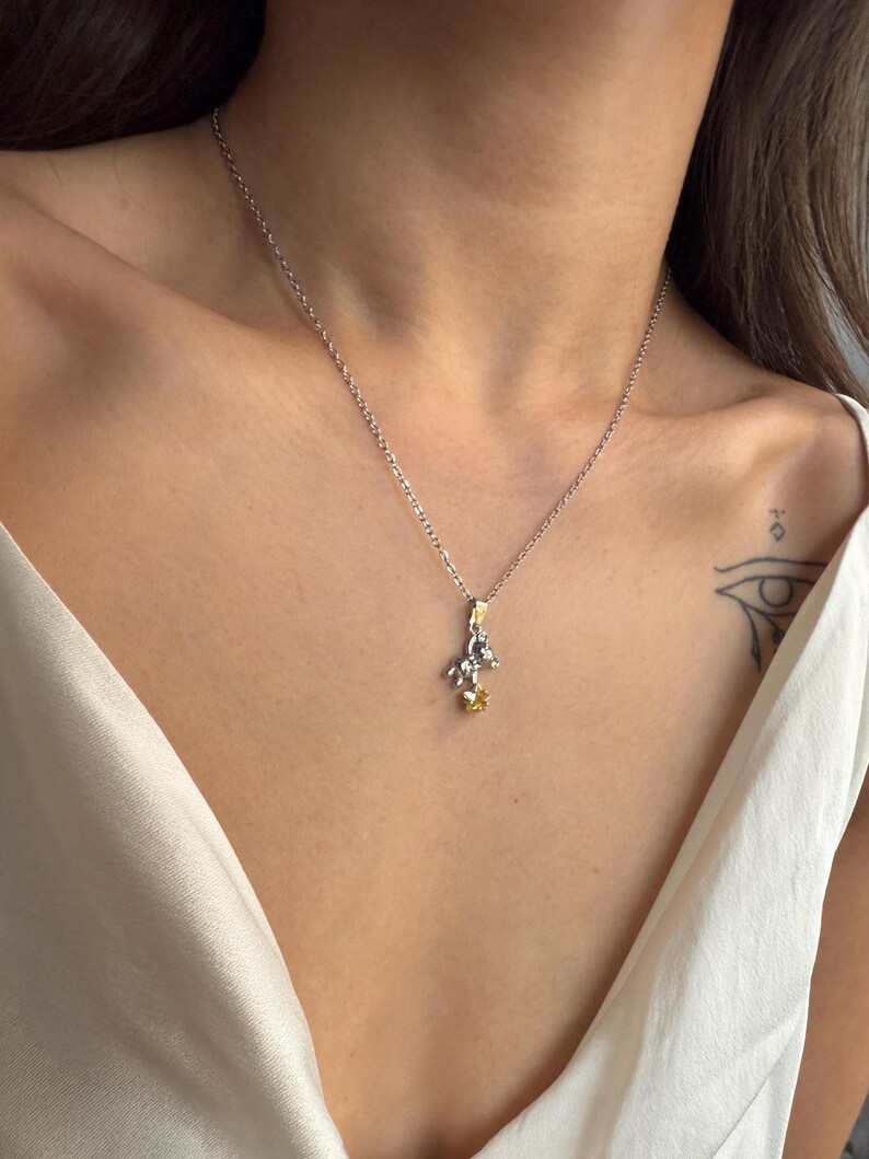May include: A silver necklace with a small pendant featuring a detailed design with gold and yellow accents. The necklace is worn on a person's chest, against a white top.