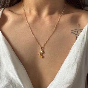 May include: A gold-colored necklace with a pendant featuring a mushroom and a yellow flower. The chain is delicate, and the pendant is centered on a white top. The necklace is a piece of jewelry.