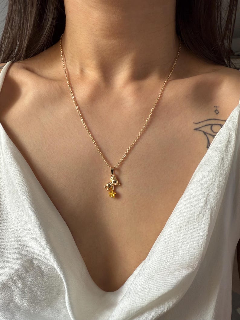May include: A gold-colored necklace with a pendant featuring a mushroom and a yellow flower. The chain is delicate, and the pendant is centered on a white top. The necklace is a piece of jewelry.