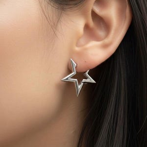 Könnte beinhalten: Ein silberner sternförmiger Ohrring. Der Ohrring ist ein Stecker, mit einer ausgehöhlten Mitte und spitzen Kanten. Der Ohrring wird am Ohr getragen, wobei Ohr und Haare im Hintergrund sichtbar sind.