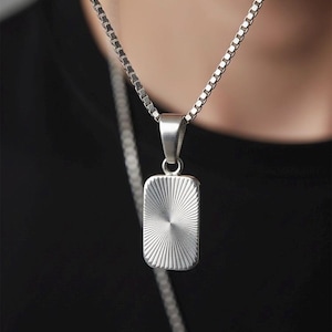 Pode incluir: Colar com pingente retangular prateado. O pingente apresenta um design texturizado de raios de sol. A corrente é do estilo elo quadrado. O colar é usado sobre uma camisa preta.