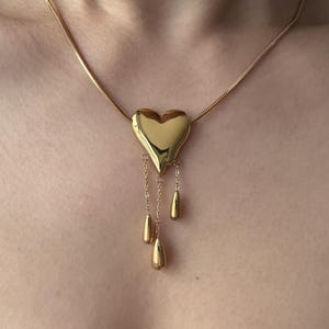 May include: A gold-colored necklace featuring a heart-shaped pendant. The pendant has three teardrop-shaped dangles suspended from delicate chains. The necklace chain is a thin, textured design.