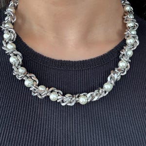 May include: A silver-toned necklace featuring a chain design interspersed with faux pearls. The necklace is worn around the neck, showcasing its intricate links and the luminous quality of the pearls. The background is a dark, solid color.