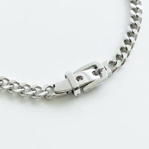 Flat lay of the silver Cuban link chain and buckle closure on a white surface, highlighting the heavyweight industrial design.