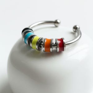 Op de afbeelding: Een zilverkleurige armband met een reeks emaille ringen in regenboogkleuren. De armband heeft een gebogen ontwerp met een bal aan elk uiteinde. De kleuren zijn onder meer blauw, geel, oranje en rood.