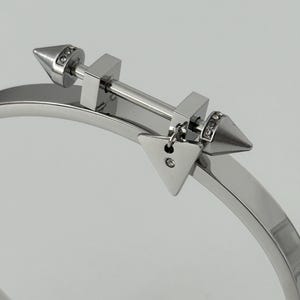 May include: A silver bangle bracelet with an arrow-themed design. The bracelet has a central bar with square features and cone-shaped ends, each embellished with tiny, clear crystals. A small, triangular charm with a single crystal hangs from the bar.