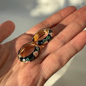 Puede incluir: Un par de pendientes de aro dorados con un diseño floral de esmalte negro. Los pendientes presentan flores rosas y blancas con hojas verdes. Los aros se sostienen en una mano sobre un fondo suave y neutro.