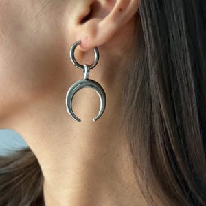 Pode incluir: Um brinco de tom prateado com um pingente de meia-lua polido suspenso por um aro circular. A forma de meia-lua está orientada com as pontas voltadas para cima, criando uma superfície metálica lisa e reflexiva.