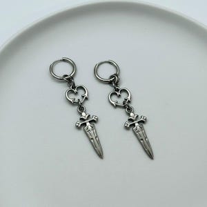 Könnte beinhalten: Ein Paar silberfarbene Ohrringe. Jeder Ohrring hat einen kleinen Reifen, ein herzförmiges Design und einen Dolch-Anhänger. Die Ohrringe werden auf einer weißen Oberfläche präsentiert.