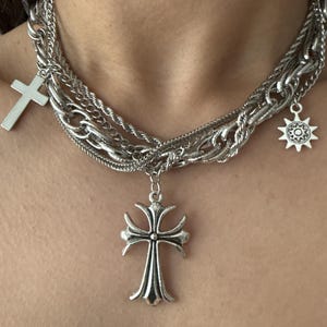 May include: Silver-toned necklace featuring multiple chains of varying styles. The necklace includes a cross pendant, a sunburst charm, and a larger decorative cross pendant. The jewelry is designed to be worn around the neck.