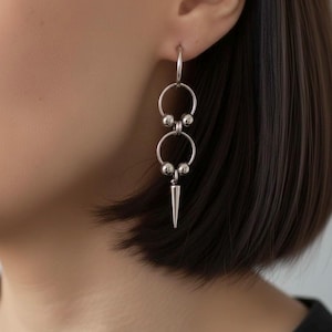 May include: Silver drop earrings with a design of linked hoops and spheres, ending in a pointed, conical charm. The earrings are displayed against a blurred backdrop, emphasizing the jewellery's design and texture.