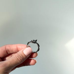 Puede incluir: Un anillo plateado con un diseño de alambre de púas. El anillo se sostiene sobre un fondo gris claro. El diseño del anillo presenta hebras de metal retorcidas, con pequeñas púas en la parte superior.