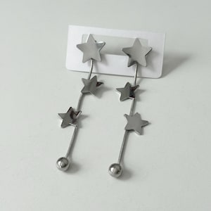 Pode incluir: Brincos pendurados prateados com um design de estrela. Cada brinco tem três estrelas descendentes e uma pequena bola na parte inferior. Os brincos estão presos a um cartão branco.