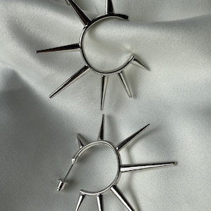 Op de afbeelding: Een paar zilverkleurige oorringen met een sunburst-ontwerp. Elke oorring heeft een cirkelvormige basis met meerdere, gelijkmatig verdeelde, puntige spikes. De oorbellen worden gepresenteerd op een witte, getextureerde ondergrond.
