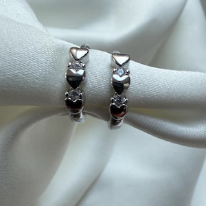 Op de afbeelding: Een paar zilveren oorringen. Elke oorring heeft drie hartvormige ontwerpen en twee heldere, ronde edelstenen. De oorbellen worden gepresenteerd op een witte, zijdeachtige stof.