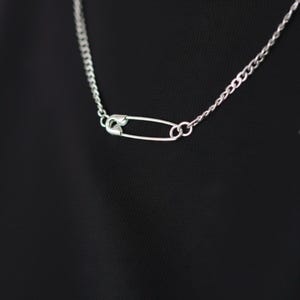 May include: Silver-toned necklace featuring a safety pin pendant. The chain is a classic curb style, and the safety pin is the focal point. The necklace is shown against a black background, highlighting the jewelry's design.